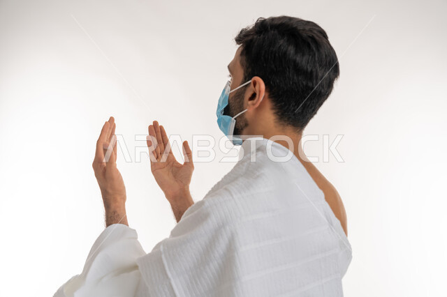 إجراءات الوقاية من فايروس كورونا، بورتريه مقرب من الجانب لشاب خليجي عربي إماراتي يرتدي ثياب الإحرام والكمامة يرفع يديه للأعلى يدعو بتضرع وخشوع، الدعاء والعبادة والتقرب إلى الله، الركن الخامس من أركان الإسلام، موسم العمرة في شهر رمضان المبارك، خلفية بيضاء