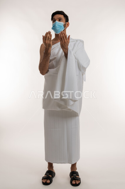 إجراءات الوقاية من فايروس كورونا، بورتريه لشاب خليجي عربي إماراتي يرتدي ثياب الإحرام والكمامة يرفع يديه للأعلى يدعو بتضرع وخشوع، الدعاء والعبادة والتقرب إلى الله، الركن الخامس من أركان الإسلام، موسم العمرة في شهر رمضان المبارك، الجسم بالطول الكامل، خلفية بيضاء
