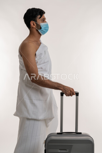 العبادة والتقرب إلى الله، الركن الخامس من أركان الإسلام، إجراءات الوقاية من فايروس كورونا، بورتريه من الجانب لشاب خليجي عربي إماراتي يرتدي ثياب الإحرام والكمامة يجر بيده حقيبة السفر، الاستعداد لمناسك الحج والعمرة، أداء الفرائض الدينية، خلفية بيضاء