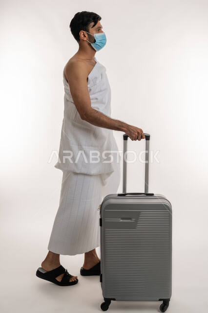 إجراءات الوقاية من فايروس كورونا، الاستعداد لإداء مناسك الحج والعمرة، بورتريه من الجانب لشاب خليجي عربي إماراتي يرتدي ثياب الإحرام والكمامة يجر بيده حقيبة السفر، العبادة والتقرب إلى الله، الركن الخامس من أركان الإسلام، الجسم بالطول الكامل، خلفية بيضاء