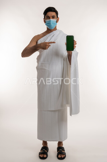 استخدام أجهزة تقنية حديثة، الاهتمام بصحة المعتمرين، اتخاذ اجراءات الوقاية من فايروس كورونا، بورتريه لشاب خليجي عربي إماراتي يرتدي ثياب الإحرام والكمامة يمسك الهاتف المحمول يشير إليه ينظر إلى الكاميرا، شاشة خضراء فارغة، الجسم بالطول الكامل، كروما، خلفية بيضاء