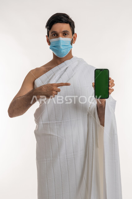Caring for the health of pilgrims, taking preventive measures against the Corona virus, portrait of a young Gulf Arab Emirati man wearing Ihram clothes and a mask holding a mobile phone pointing at it and looking at the camera, using modern technical devices, empty green screen, chroma key, white background