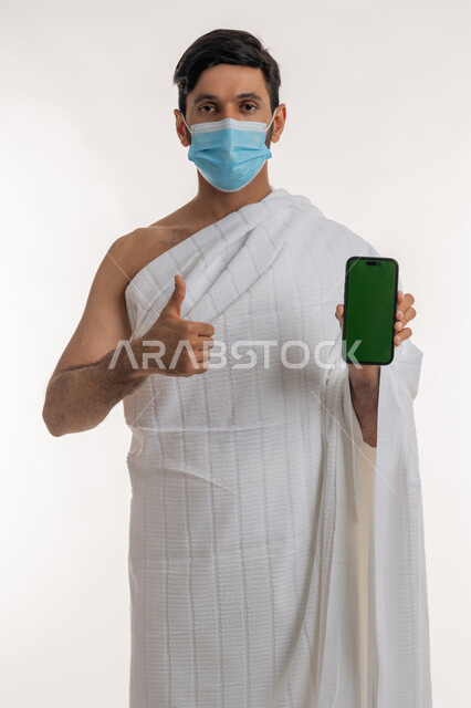 اتخاذ إجراءات الوقاية من فايروس كورونا، بورتريه لشاب خليجي عربي إماراتي يرتدي ثياب الإحرام والكمامة يمسك الهاتف المحمول يشير بإبهامه للأعلى، استخدام الأجهزة التقنية الحديثة، تعابير الجودة والامتياز، شاشة خضراء فارغة، كروما، خلفية بيضاء