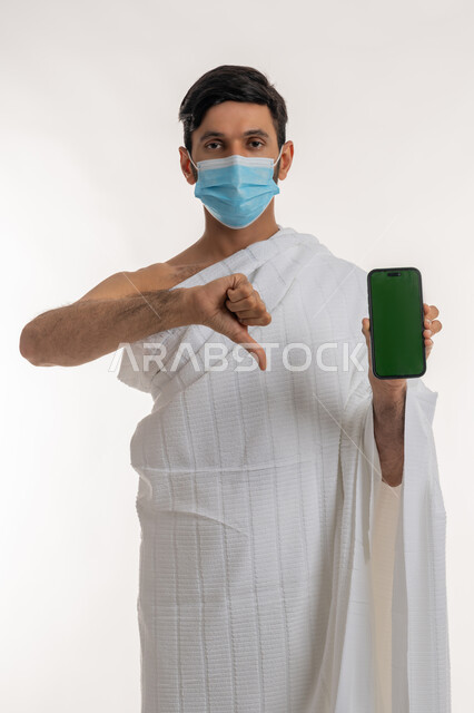 اتخاذ اجراءات الوقاية من فايروس كورونا، بورتريه لشاب خليجي عربي إماراتي يرتدي ثياب الإحرام والكمامة يمسك الهاتف المحمول يشير بإبهامه للإسفل بتعابير الرفض وعدم الإعجاب، استخدام الأجهزة التقنية الحديثة، شاشة خضراء فارغة، كروما، خلفية بيضاء