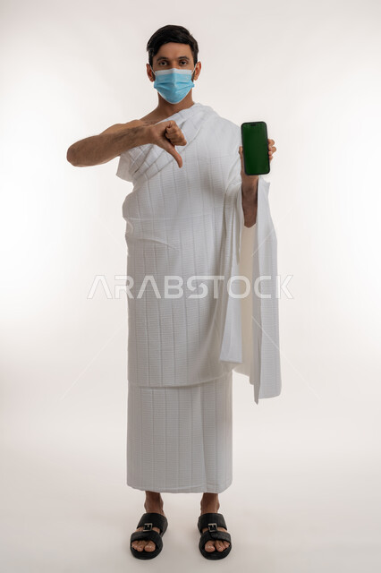 استخدام الأجهزة التقنية الحديثة، بورتريه لشاب خليجي عربي إماراتي يرتدي ثياب الإحرام والكمامة يمسك الهاتف المحمول يشير بإبهامه للإسفل بتعابير الرفض وعدم الإعجاب، اتخاذ اجراءات الوقاية من فايروس كورونا، شاشة خضراء فارغة، الجسم بالطول الكامل، كروما، خلفية بيضاء