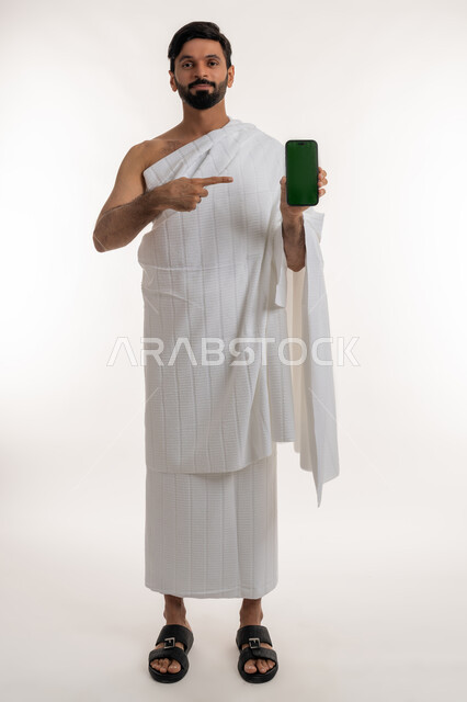 عرض تطبيقات وبرامج دليل الحاج، بورتريه لشاب خليجي عربي إماراتي يرتدي ثياب الإحرام  يمسك الهاتف المحمول يشير إليه ينظر إلى الكاميرا بإيماءات السرور، استخدام الأجهزة التقنية الحديثة، شاشة خضراء فارغة، الجسم بالطول الكامل، كروما، خلفية بيضاء