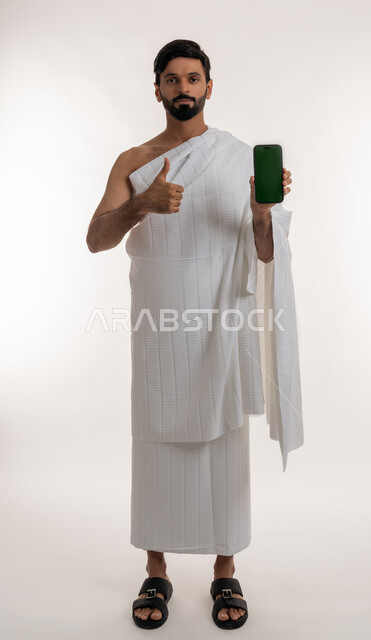 عرض تطبيقات وبرامج دليل الحاج، تعابير الجودة والامتياز، بورتريه لشاب خليجي عربي إماراتي يرتدي ثياب الإحرام  يمسك الهاتف المحمول يشير بإبهامه للأعلى ينظر إلى الكاميرا، استخدام الأجهزة التقنية الحديثة، شاشة خضراء فارغة، الجسم بالطول الكامل، كروما، خلفية بيضاء