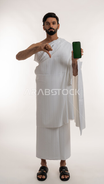 استخدام الأجهزة التقنية الحديثة، بورتريه لشاب خليجي عربي إماراتي يرتدي ثياب الإحرام يمسك الهاتف المحمول ينظر إلى الكاميرا يشير بإبهامه للإسفل بتعابير الرفض وعدم الإعجاب، عرض تطبيقات دليل الحاج والمعتمر، شاشة خضراء فارغة، الجسم بالطول الكامل، كروما، خلفية بيضاء