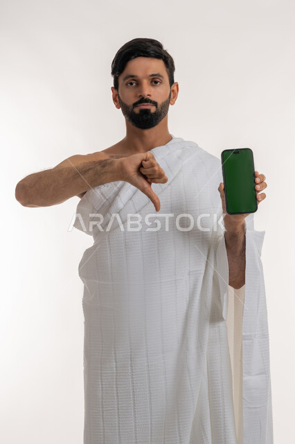عرض تطبيقات دليل الحاج والمعتمر على الجوال، بورتريه لشاب خليجي عربي إماراتي يرتدي ثياب الإحرام يحمل بيده الهاتف المحمول ينظر إلى الكاميرا يشير بإبهامه للإسفل بتعابير الرفض وعدم الإعجاب، استخدام الأجهزة التقنية الحديثة، شاشة خضراء فارغة، كروما، خلفية بيضاء