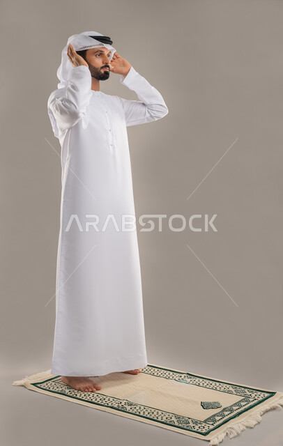 Performing prayers on time, worship and getting closer to Allah, side portrait of an Arab Gulf Emirati man wearing a kandura and ghutra standing straight performing the obligatory prayer raising his hands for the Takbirat al-Ihram, concept of the pillars of Islam, feeling of humility and tranquility, full-length body image, gray background