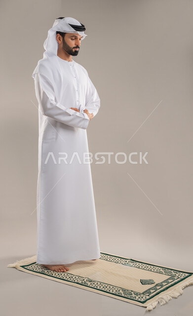 الشعور بالخشوع والطمأنينة والسكون، أداء الصلوات على وقتها، العبادة والتقرب إلى الله، بورتريه من الجانب لرجل عربي خليجي إماراتي يرتدي الكندورة والغترة يقف باستقامة على السجادة يؤدي فريضة الصلاة، الركن الثاني من أركان الإسلام، صورة الجسم بالطول الكامل، خلفية رمادية