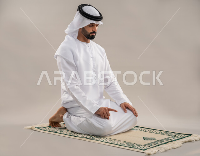 أداء الصلوات على وقتها، العبادة والتقرب إلى الله، بورتريه من الجانب لرجل عربي خليجي إماراتي يرتدي الكندورة والغترة يجلس على سجادة الصلاة يقوم بالتشهد، الشعور بالخشوع والطمأنينة والسكون، الركن الثاني من أركان الإسلام، خلفية رمادية
