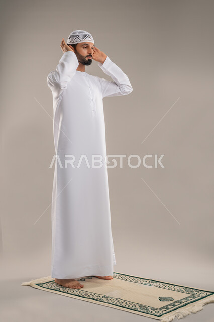 Humility, supplication and beseeching Allah, concept of worship and observing prayers, side portrait of an Emirati Arab man wearing a kandura and white cap standing straight raising his hands for Takbirat al-Ihram performing the obligatory prayer, the second pillar of Islam, full-length body image, gray background