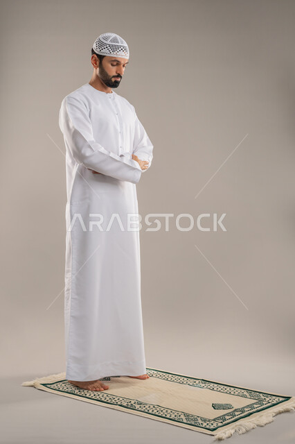 مفهوم العبادة والحفاظ على الصلوات، الخشوع والتوسل والتضرع لله، بورتريه من الجانب لرجل عربي خليجي إماراتي يرتدي الكندورة وطاقية بيضاء يقف باستقامة على السجادة يؤدي فريضة الصلاة، الركن الثاني من أركان الإسلام، صورة الجسم بالطول الكامل، خلفية رمادية