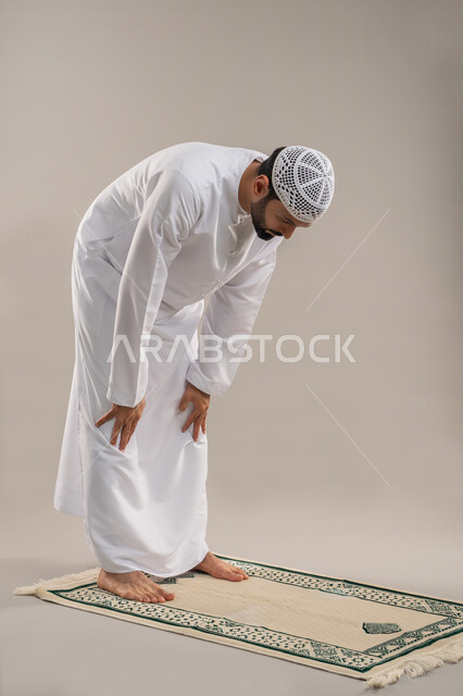 أداء الصلوات على وقتها، بورتريه من الجانب لرجل عربي خليجي إماراتي يرتدي الكندورة وطاقية بيضاء يركع على سجادة الصلاة بإيماءات الخشوع والتوسل والتضرع، التقرب الى الله والاجتهاد في العبادة، خلفية رمادية