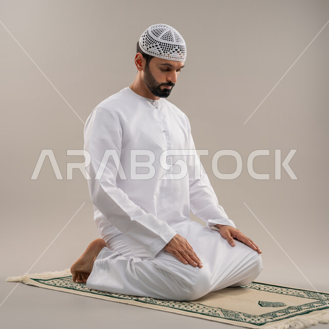 الركن الثاني من أركان الإسلام، الخشوع والتوسل والتضرع لله والاجتهاد في العبادة، بورتريه من الجانب لرجل عربي خليجي إماراتي يرتدي الكندورة وطاقية بيضاء يجلس على السجادة يؤدي فريضة الصلاة، خلفية رمادية