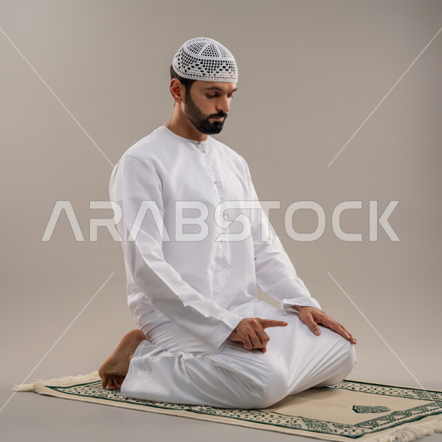 الركن الثاني من أركان الإسلام، الخشوع والتوسل والتضرع لله والاجتهاد في العبادة، بورتريه من الجانب لرجل عربي خليجي إماراتي يرتدي الكندورة وطاقية بيضاء يجلس على السجادة يؤدي فريضة الصلاة، خلفية رمادية