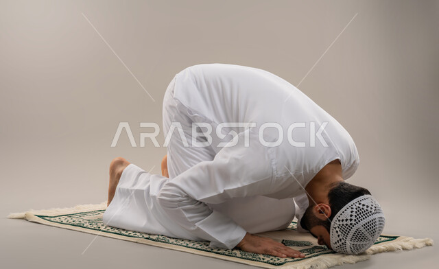 Getting closer to God and striving in worship, performing prayers on time, side portrait of an Arab Gulf Emirati man wearing a kandura and a white cap, prostrating on a prayer rug with gestures of reverence, supplication and entreaty, gray background