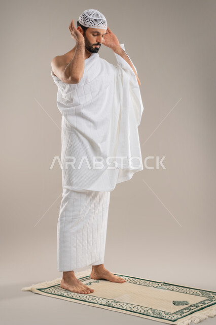 أداء مناسك الحج والعمرة، مفهوم العبادة والحفاظ على الصلوات، بورتريه من الجانب لرجل عربي خليجي إماراتي يرتدي ثياب الإحرام وطاقية بيضاء يقف باستقامة يرفع يديه لتكبيرة الإحرام يؤدي فريضة الصلاة، الركن الثاني من أركان الإسلام، صورة الجسم بالطول الكامل، خلفية رمادية