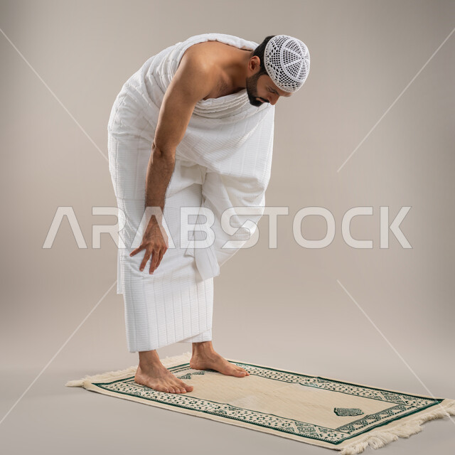التقرب الى الله من خلال الاجتهاد في العبادة وتأدية مناسك الحج والعمرة، أداء الصلوات على وقتها، بورتريه من الجانب لرجل عربي خليجي إماراتي يرتدي ثياب الإحرام وطاقية بيضاء يركع على سجادة الصلاة بإيماءات الخشوع والتوسل والتضرع، خلفية رمادية