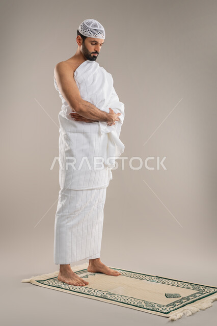 The second pillar of Islam, humility, supplication, and supplication to Allah and diligence in worship, side portrait of an Arab Gulf Emirati man wearing Ihram clothes and a white cap standing straight on the prayer mat performing the ritual of Hajj and Umrah, full body image, gray background