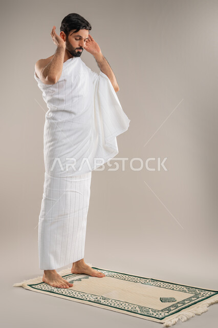 Performing Hajj and Umrah rituals, concept of worship and maintaining prayers, side portrait of an Arab Gulf Emirati man wearing Ihram clothes standing straight raising his hands for Takbeer Al-Ihram performing the obligatory prayer, the second pillar of Islam, full body image, gray background