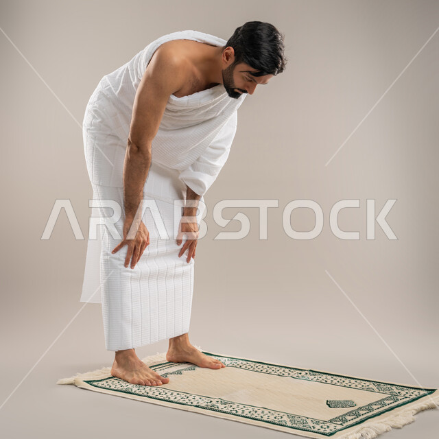 Performing prayers on time, getting closer to God through diligence in worship and performing the Hajj and Umrah rituals, side portrait of an Arab Gulf Emirati man wearing Ihram clothes kneeling on a prayer rug with gestures of reverence, supplication and entreaty, gray background