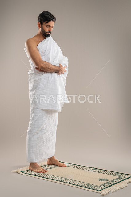 The second pillar of Islam, humility, supplication, and supplication to Allah and diligence in worship, side portrait of an Arab Gulf Emirati man wearing Ihram clothes standing straight on the prayer mat performing the obligatory prayer, performing the rituals of Hajj and Umrah, full body image, gray background