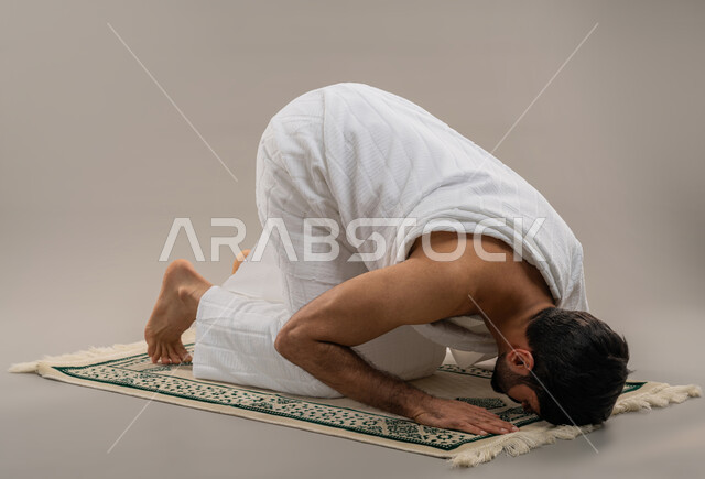 أداء الصلوات على وقتها، مناسك الحج والعمرة، التقرب الى الله والاجتهاد في العبادة، بورتريه من الجانب لرجل عربي خليجي إماراتي يرتدي ثياب الإحرام يسجد على سجادة الصلاة بإيماءات الخشوع والتوسل والتضرع، خلفية رمادية