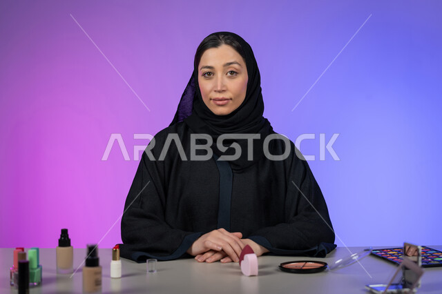 الاهتمام بالمظهر الخارجي، مفهوم الأنوثة والنعومة، بورتريه لامرأة عربية خليجية اماراتية ترتدي العباية والحجاب تضع يدها فوق بعضهم البعض وأمامها مجموعة من أدوات ومستحضرات التجميل، النظر إلى الكاميرا بتعابير السرور، شابة سعودية أنيقة، خلفية ملونة