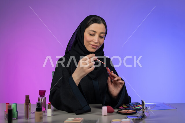 الشغف بوضع الميكاب, امرأة سعودية خبيرة بمستحضرات التجميل، مفهوم الإهتمام بالجمال والبشرة، التسويق لمنتجات ومستحضرات الجمال في المتاجر و عبر الإنترنت، بورتريه مقرب لشابة عربية خليجية امارتية محجبة تضع قلم أحمر الشفاه، خلفية ملونة