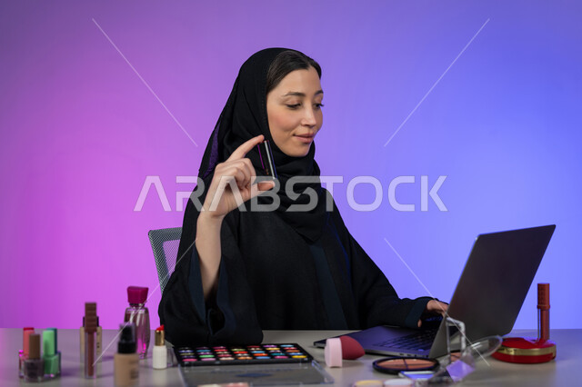ترويج منتجات وأدوات المكياج عبر الانترنت، بورتريه لسيدة عربية خليجية اماراتية محجبة ترتدي العباءة السوداء تسوق لبيع مستحضرات التجميل باستخدام الحاسب المحمول، الإهتمام بالجمال والمظهر الخارجي، شابة سعودية تعرض أدوات مكيآب تتوريال ، خلفية ملونة