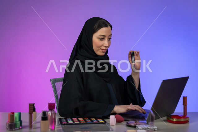 Beauty and appearance interest, promoting makeup products and tools online, portrait of a veiled Arab Gulf Emirati woman wearing a black abaya shopping for cosmetics using a laptop, a young Saudi woman displaying makeup tutorial tools, colorful background