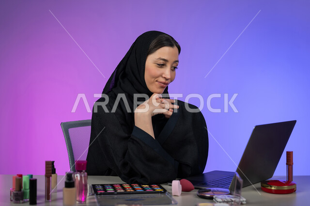 الأنوثة والاهتمام بالجمال والبشرة والمظهر الخارجي, خبيرة سعودية بمستحضرات التجميل النسائية، بورتريه مقرب لإمرأة عربية خليجية اماراتية ترتدي العباءة والحجاب تنظر إلى الحاسب المحمول وتضع يدها على ذقنها بايماءات السرور والسعادة، التسويق لمنتجات الميكآب، خلفية ملونة