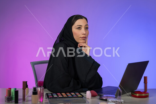 مفهوم الأنوثة والنعومة، الاهتمام بالمظهر الخارجي، بورتريه لامرأة عربية خليجية اماراتية ترتدي العباءة السوداء والحجاب تضع يدها على ذقنها بإيماءات التفكير والتخطيط للمستقبل، النظر إلى شيء ما، التسويق لمنتجات وأدوات التجميل عبر الانترنت، شابة سعودية أنيقة، خلفية ملونة