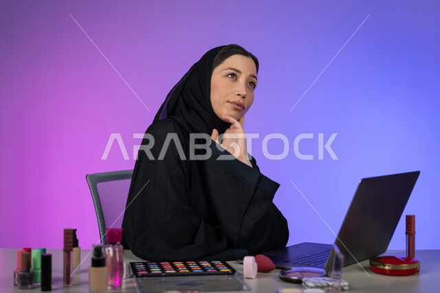 مفهوم الأنوثة والنعومة، الاهتمام بالمظهر الخارجي، بورتريه لامرأة عربية خليجية اماراتية ترتدي العباءة السوداء والحجاب تضع يدها على ذقنها بإيماءات التفكير والتخطيط للمستقبل، النظر إلى شيء ما، التسويق لمنتجات وأدوات التجميل عبر الانترنت، شابة سعودية أنيقة، خلفية ملونة