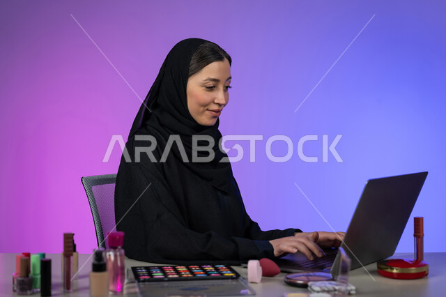 مفهوم التسويق عبر الانترنت، الترويج لمنتجات ومستحضرات التجميل عبر المواقع الالكترونية، إستخدام جهاز الحاسوب المحمول، بورتريه لإمراة عربية خليجية اماراتية محجبة ترتدي العباءة السوداء خبيرة في الميكآب، شابة سعودية أنيقة، خلفية ملونة