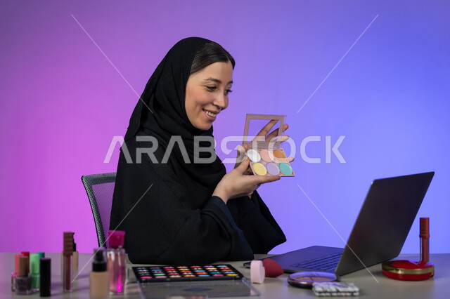 Online promotion of makeup products and tools, portrait of a veiled Arab Gulf Emirati woman wearing a black abaya shopping for cosmetics using a laptop, interest in beauty and appearance, a young Saudi woman displaying makeup tutorials, colorful background
