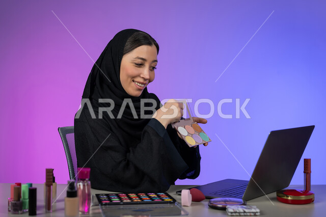Online promotion of makeup products and tools, portrait of a veiled Arab Gulf Emirati woman wearing a black abaya shopping for cosmetics using a laptop, interest in beauty and appearance, a young Saudi woman displaying makeup tutorials, colorful background