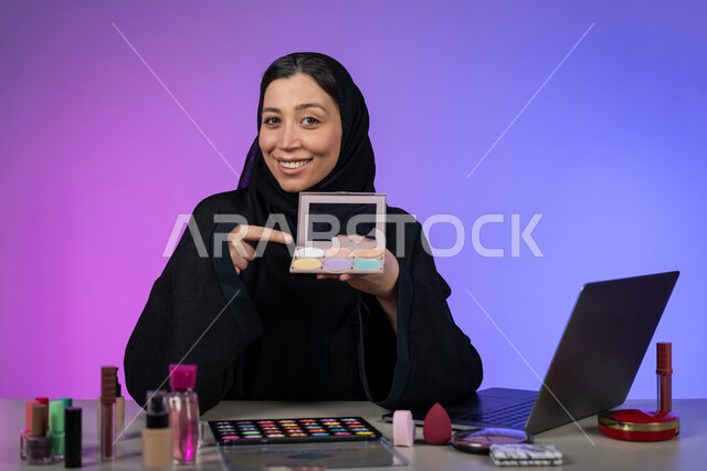 مفهوم التسويق عبر الانترنت، الترويج لمنتجات ومستحضرات التجميل عبر المواقع الالكترونية، إستخدام جهاز الحاسوب المحمول، بورتريه لإمراة عربية خليجية اماراتية محجبة ترتدي العباءة السوداء خبيرة في الميكآب، شابة سعودية أنيقة، خلفية ملونة