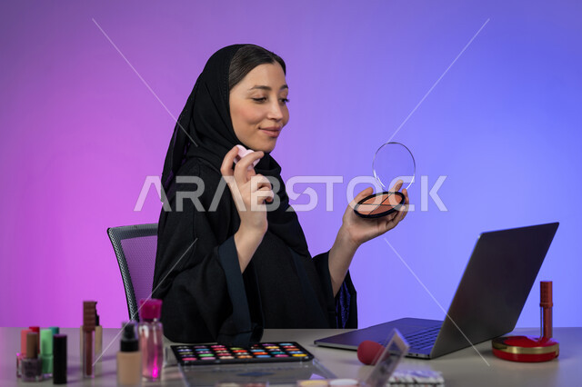 ترويج منتجات وأدوات المكياج عبر الانترنت، بورتريه لسيدة عربية خليجية اماراتية محجبة ترتدي العباءة السوداء تسوق لبيع مستحضرات التجميل باستخدام الحاسب المحمول، الإهتمام بالجمال والمظهر الخارجي، شابة سعودية تعرض أدوات مكيآب تتوريال ، خلفية ملونة