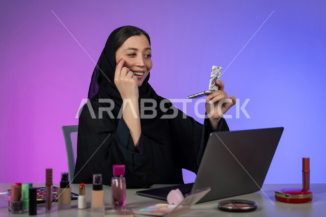 الإهتمام بالجمال والمظهر الخارجي، ترويج منتجات وأدوات المكياج عبر الانترنت، بورتريه لسيدة عربية خليجية اماراتية محجبة ترتدي العباءة السوداء تسوق لبيع مستحضرات التجميل باستخدام الحاسب المحمول، شابة سعودية تعرض أدوات مكيآب تتوريال ، خلفية ملونة