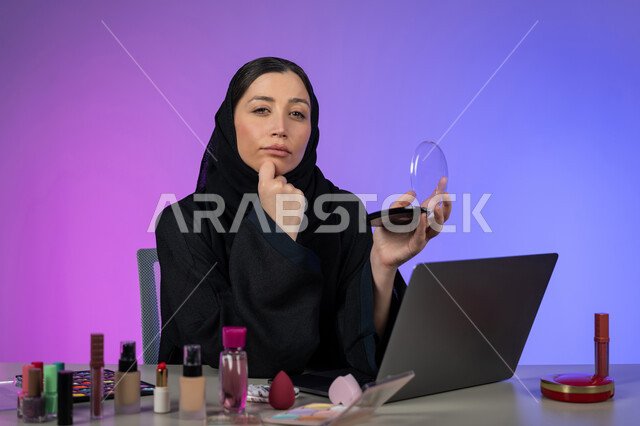 النظر بشرود إلى مكان ما, التمعن والتركيز والتخطيط للمستقبل، بورتريه مقرب لامرأة عربية خليجية اماراتية محجبة تضع يدها على ذقنها وتنظر إلى الكاميرا بتفكير وتأمل، شابة سعودية خبيرة تجميل تفكر بتسويق منتجات و مساحيق التجميل عبر الانترنت، خلفية ملونة