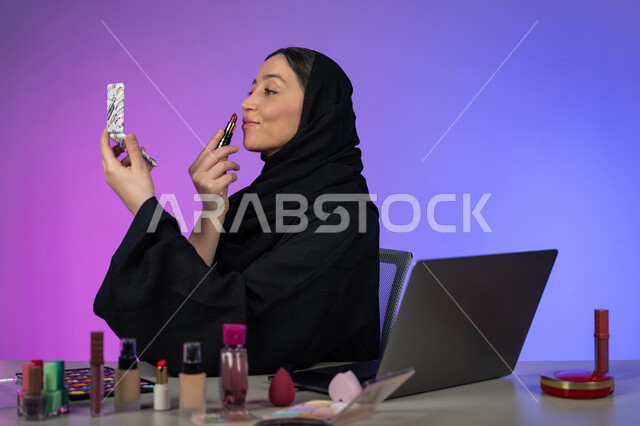 التجمل والتزين بادوات التجميل, التسويق لمنتجات ومستحضرات المكياج عبر الإنترنت، مفهوم الإهتمام بالجمال والبشرة، بورتريه لخبيرة تجميل عربية خليجية اماراتية محجبة ترتدي العباءة السوداء تقوم بوضع الميكآب وتنظر إلى المرآة، شابة سعودية تستعمل أدوات مكياج تتوريال