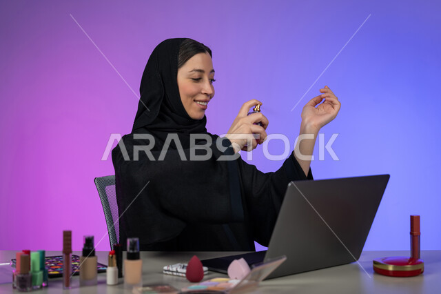 التسويق لمنتجات الروائح الوطنية الطيبة و الفاخرة، مفهوم الأناقة والاهتمام بالمظهر الخارجي، استخدام الزيوت العطرية الطبيعية، بورتريه لسيدة عربية خليجية إماراتية محجبة ترتدي عباءة سوداء تمسك بيدها زجاجة وتقوم برش العطر, شابة سعودية أنيقة تروج للعطور، خلفية ملونة