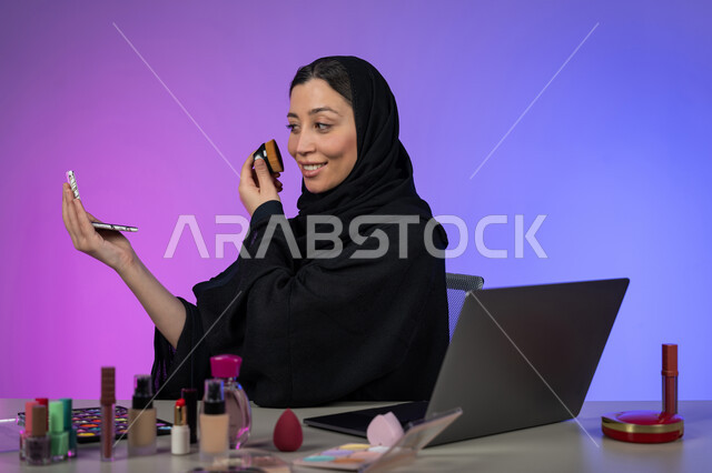 التجمل والتزين بادوات التجميل, التسويق لمنتجات ومستحضرات المكياج عبر الإنترنت، مفهوم الإهتمام بالجمال والبشرة، بورتريه لخبيرة تجميل عربية خليجية اماراتية محجبة ترتدي العباءة السوداء تقوم بوضع الميكآب وتنظر إلى المرآة، شابة سعودية تستعمل أدوات مكياج تتوريال