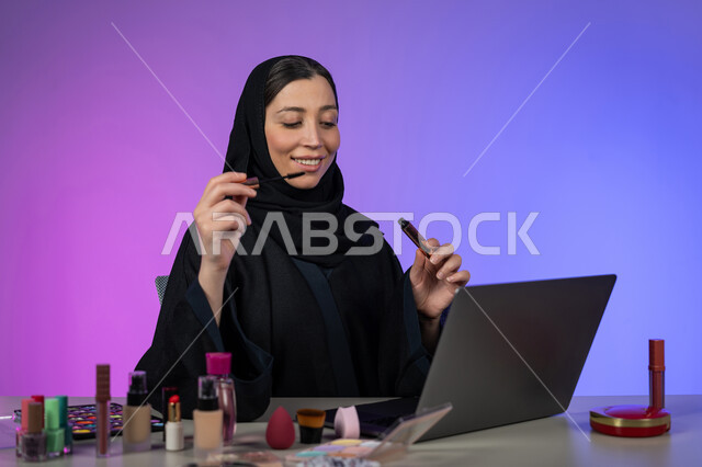 التسويق لمنتجات ومستحضرات الميكآب عبر الإنترنت، الشغف بوضع المكياج, مفهوم الإهتمام بالجمال والبشرة، بورتريه لخبيرة تجميل عربية خليجية اماراتية محجبة ترتدي العباءة السوداء تضع المسكرة لإبراز العين، شابة سعودية تعرض منتجاتها للبيع، خلفية ملونة