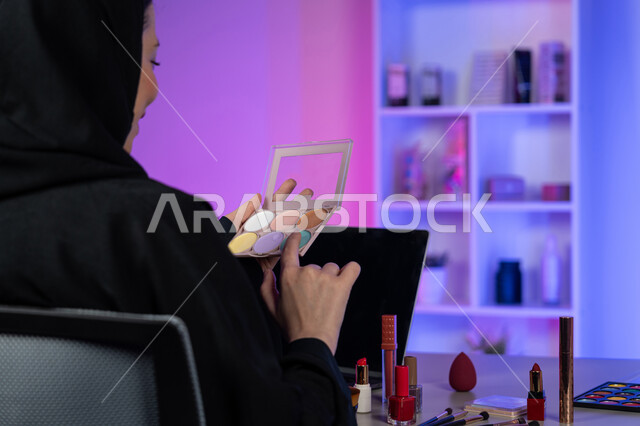 التسويق لمنتجات التجميل، متجر بيع مستحضرات وأدوات المكياج، مفهوم الإهتمام بالجمال والبشرة، صورة من الخلف لإمرأة عربية خليجية اماراتية محجبة ترتدي العباءة السوداء تحمل بيدها باليت ظل العيون الملونة، خبيرة تجميل سعودية