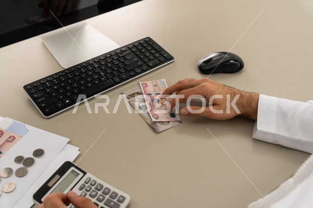 السعي نحو التوفير والادخار، محاولة إدارة المصروف المنزلي بتدبر، استخدام الحاسبة الرقمية في الأعمال الاقتصادية، صورة مقربة من الخلف لرجل عربي خليجي اماراتي يرتدي الكندورة والغترة البيضاء يجلس على كرسي مريح يقوم بالحسابات الشهرية المالية