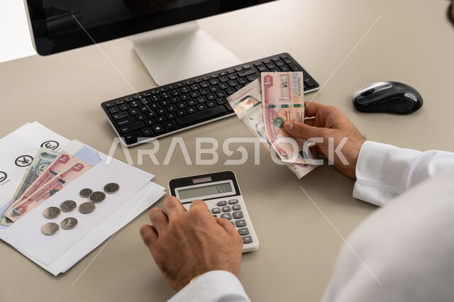 استخدام الحاسبة الرقمية في الأعمال الاقتصادية، محاولة إدارة المصروف المنزلي بتدبر، السعي نحو التوفير والادخار، صورة مقربة من الخلف لرجل عربي خليجي اماراتي يرتدي الكندورة والغترة البيضاء يجلس على كرسي مريح يقوم بالحسابات الشهرية المالية